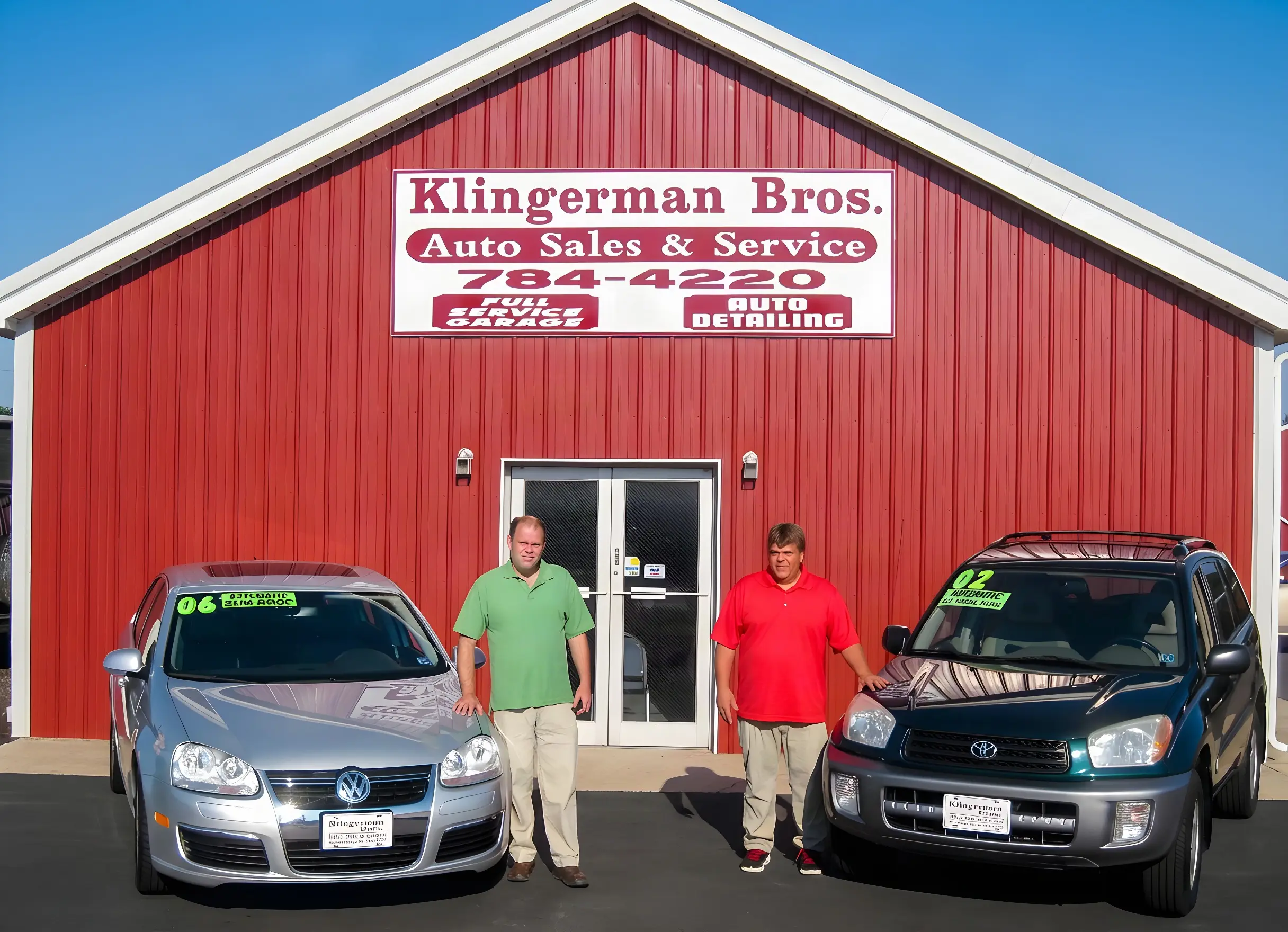 The red Klingerman Bros. shop building with two men standing between two cars out front